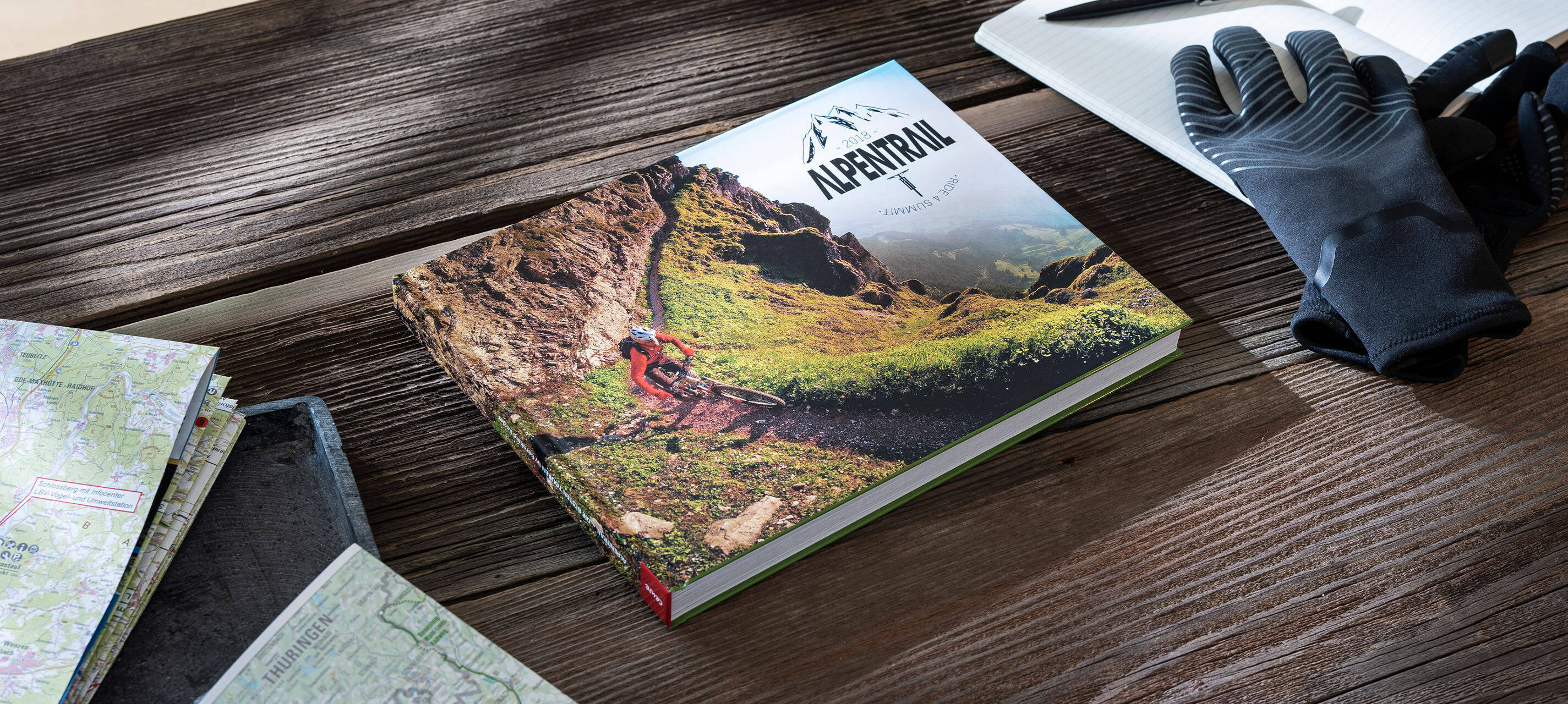 Auf einem Tisch liegen ein Fotobuch, Handschuhe, ein Heft und Landkarten. Auf dem Fotobuch sind ein Mountainbike sowie die Aufschrift “Alpentrail 2018” zu sehen.