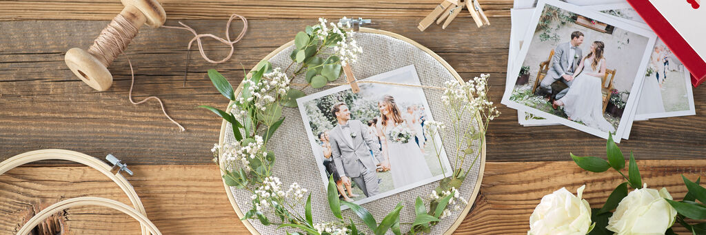 Stickkranz und Square Print mit Hochzeitsmotiv liegt mit Zweigen auf Holztisch. Daneben sind Kordel, Pflanzen, Schere und Decke platziert. Oben rechts im Bild ist eine Foto-Box mit Square Prints zu sehen.