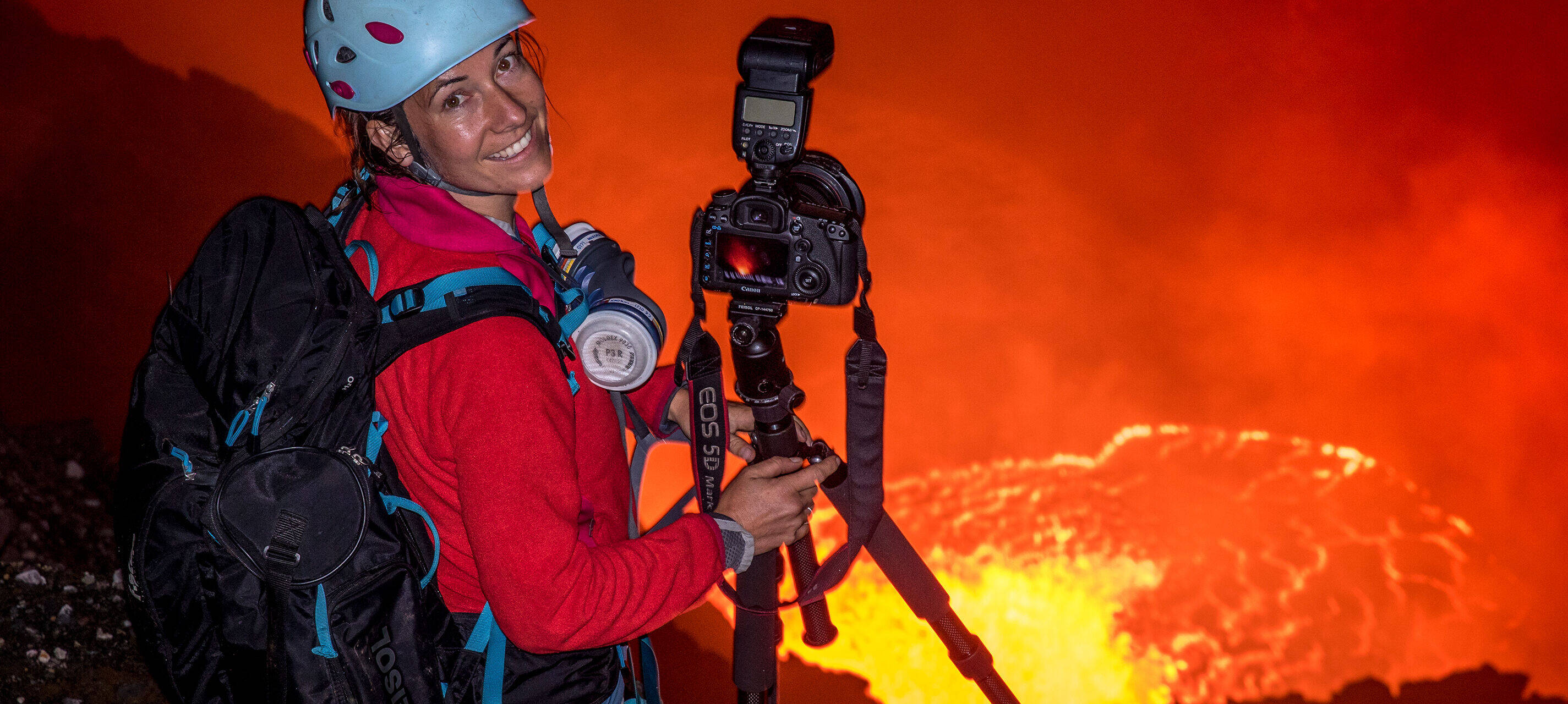 Expeditionsfotografin Ulla Lohmann steht mit Helm und Rucksack am Rande eines Vulkans und lächelt die Betrachtenden an. Neben ihr steht eine Kamera mit Stativ.