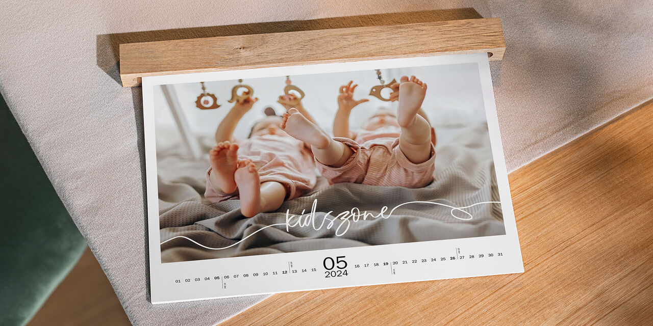 Auf einem Tisch aus Holz liegt ein Wandkalender mit Babymotiven im Querformat. Sichtbar ist das Blatt für den Monat Mai. Eine Holzleiste verdeckt die Ringbindung des Kalenders.