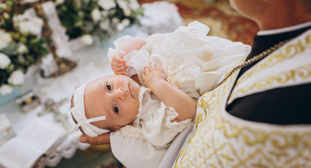 Baby in weißem Kleid und Stirnband liegt auf dem Arm einer erwachsenen Person in festlicher Kleidung.