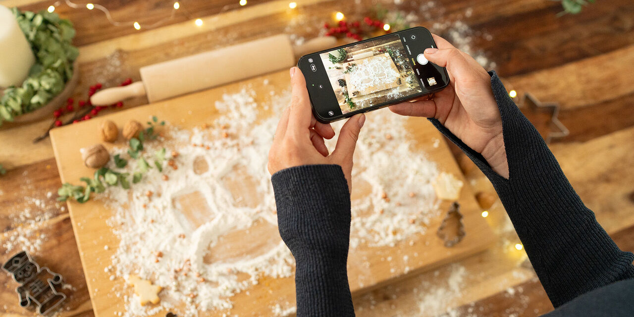 Es ist ein großes, mit Mehl bestreutes Holzbrett zu erkennen, das weihnachtlich dekoriert wurde. Im Vordergrund sind zwei Hände zu sehen, die ein Smartphone horizontal festhalten und mit diesem ein Foto von dem Brett machen.