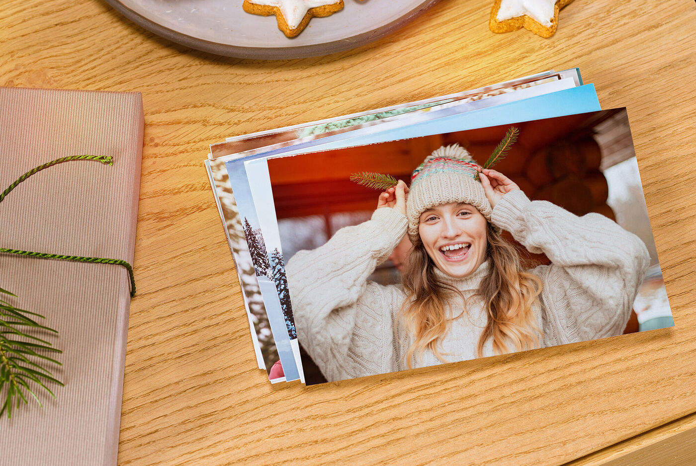 Eine fröhliche Person trägt einen weißen Pullover und eine kreative Mütze mit Hörnern, während sie sich in einer gemütlichen Umgebung befindet. Auf einem Tisch liegen verschiedene Fotografien sowie weihnachtliche Plätzchen.