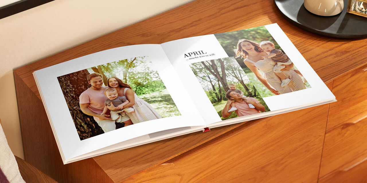 Auf einem Holzschrank liegt das aufgeklappte Fotobuch. Auf der linken Seite ist ein Familienporträt mit Vater, Mutter und Kind zu sehen. Auf der rechten Seite befinden sich zwei Bilder, auf denen jeweils Mutter und Vater mit dem Kind abgebildet sind. Darüber steht die Überschrift „April“.