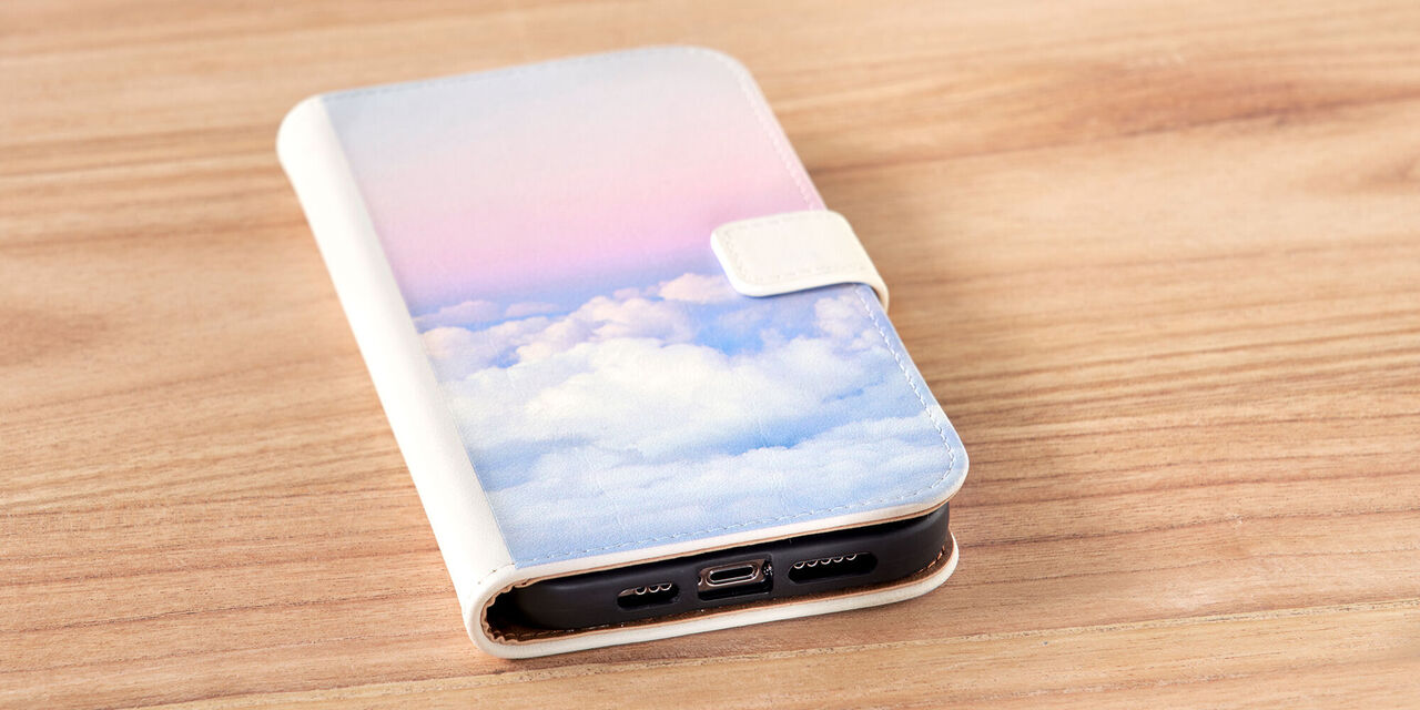 Auf einem Tisch liegt ein Smartphone. Es ist die Handyhülle zu sehen, auf der eine Aufnahme von rosafarbenen Wolken zu sehen ist. Bei der Handyhülle handelt es sich um eine Sideflip Tasche aus weißem Leder.
