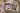 Auf einem Sofa liegt ein geöffnetes CEWE FOTOBUCH neben einem Kuscheltier und einer Decke. Auf der linken Seite ist das Foto einer Hand zu erkennen, die eine Kinderzeichnung von einer Familie festhält. Die Überschrift lautet „Wir fünf“. Auf der rechten Seite ist ein Familienfoto mit den Eltern und drei Kindern zu sehen. Der Hintergrund der Doppelseite ist mit einem violetten Aquarellfarbverlauf gestaltet.