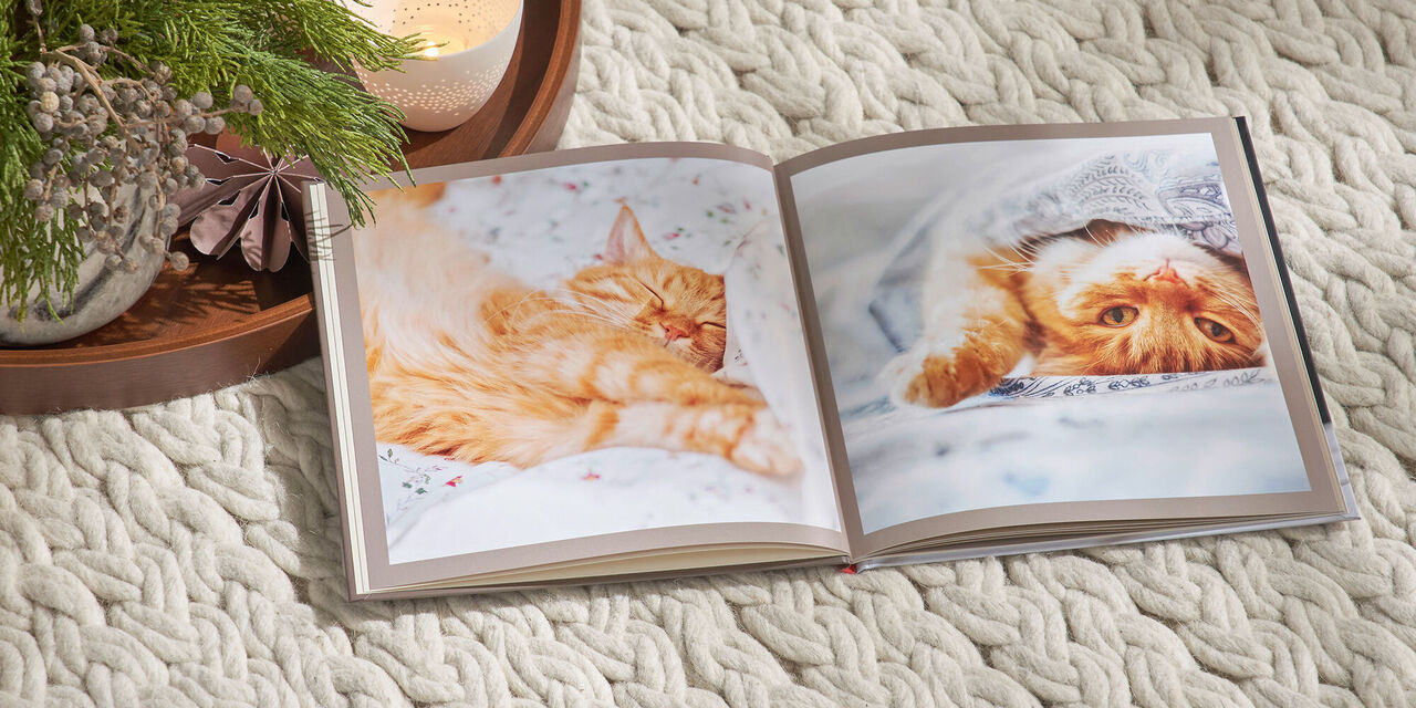 Ein aufgeschlagenes CEWE FOTOBUCH im quadratischen Format liegt auf einer weißen Decke. Auf der Doppelseite sind Fotos einer roten Katze zu sehen. Links neben dem Fotobuch liegt ein Tablett mit weihnachtlicher Deko und Kerzen.