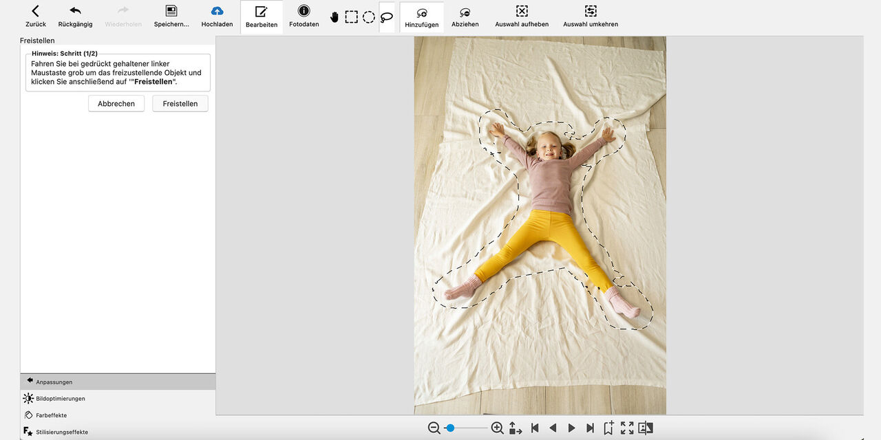 Screenshot der CEWE Fotowelt Software: Es ist ein Mädchen zu sehen, das auf dem Boden auf einer hellen, ausgebreiteten Decke liegt und in die Kamera lächelt. Beide Arme und Beine hat das Mädchen dabei weit von sich gestreckt. Die Kontur des Körpers ist grob durch eine gestrichelte Linie umrandet.