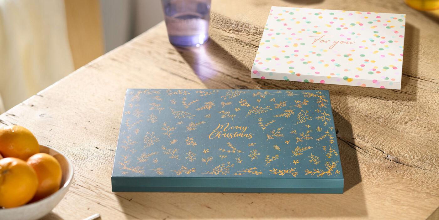 Zwei Geschenkverpackungen für das CEWE FOTOBUCH: eine in dunkelgrün mit „Merry Christmas“ und dahinter eine in weiß mit bunten Punkten liege