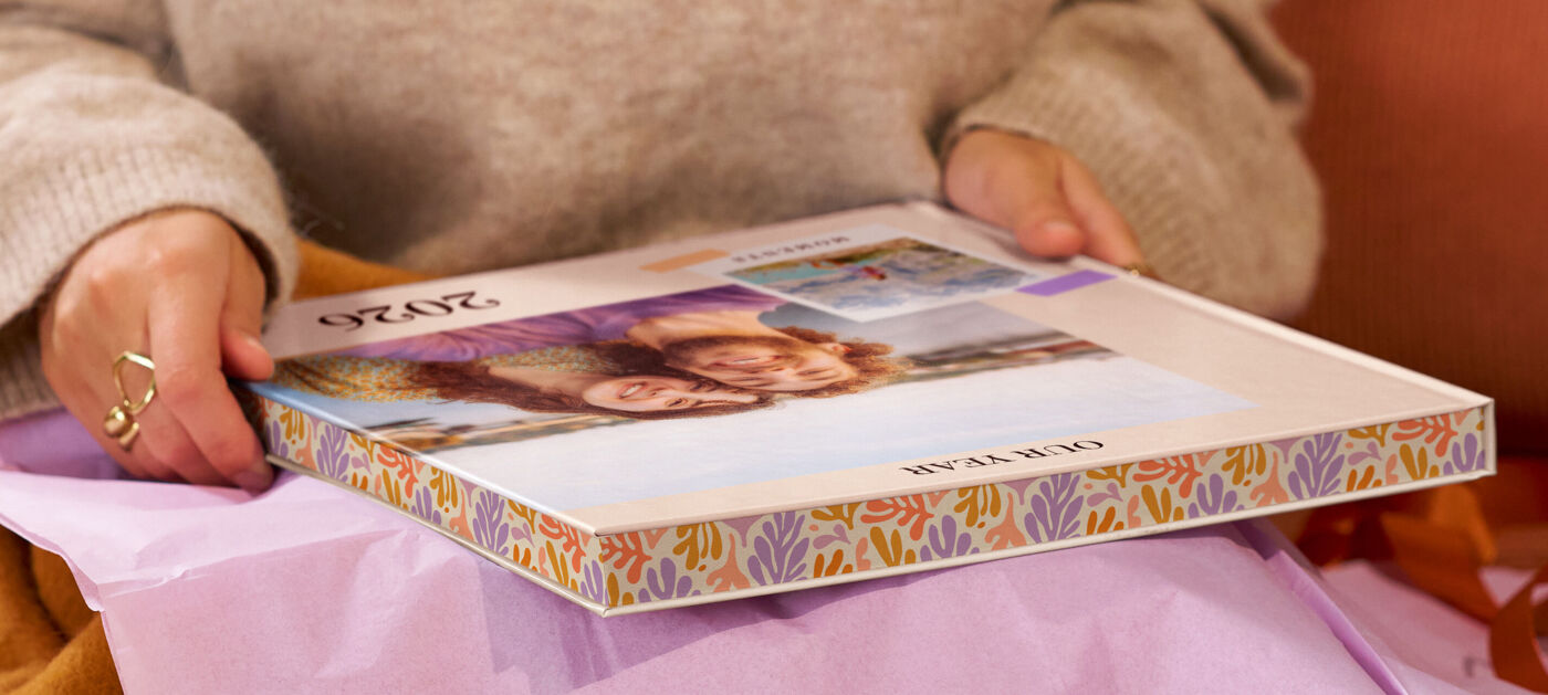 Eine Frau sitzt auf einem Sofa und packt ein Geschenk aus, in dem sich ein CEWE FOTOBUCH mit Farbschnitt befindet.