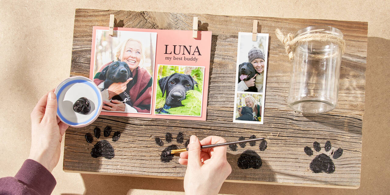 Zwei Hände malen mit schwarzer Farbe die kleinen Hundepfoten auf das Holzbrett.