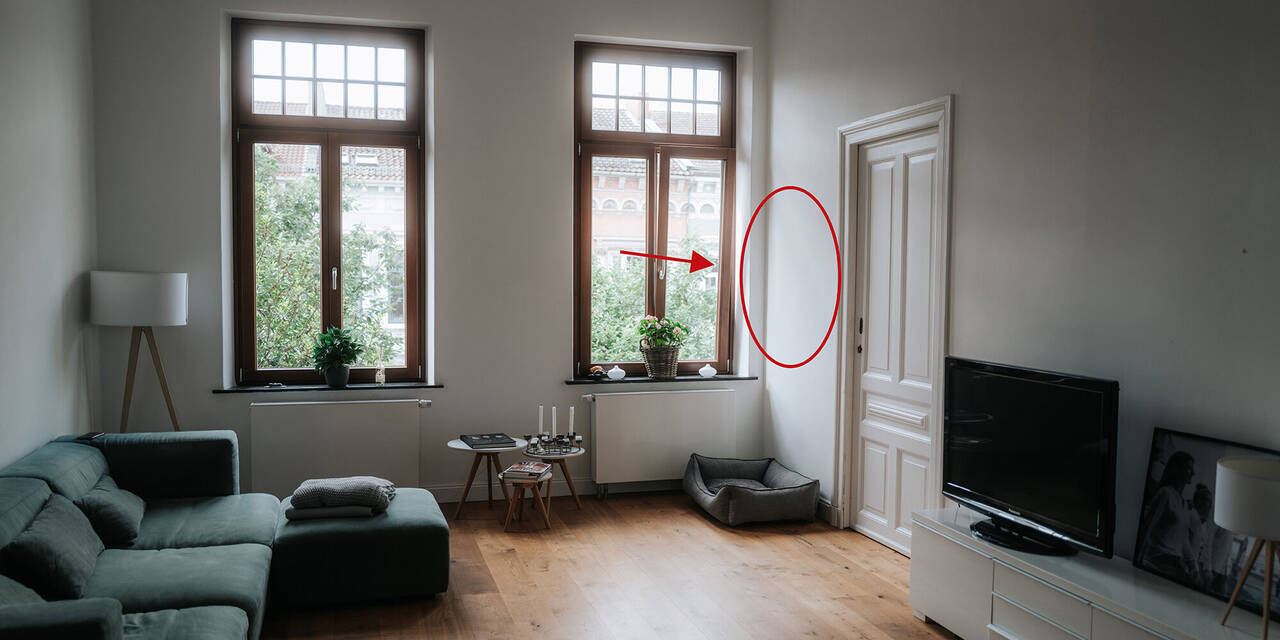 Auf dem Foto ist ein Wohnzimmer mit zwei großen Fenstern zu sehen. Auf der linken Seite steht eine grüne Couch und dahinter eine Lampe. Auf der rechten Seite befindet sich ein Sideboard mit einem Fernseher und dahinter eine Tür. Auf der rechten Seite des Fotos ist ein roter Kreis eingezeichnet.